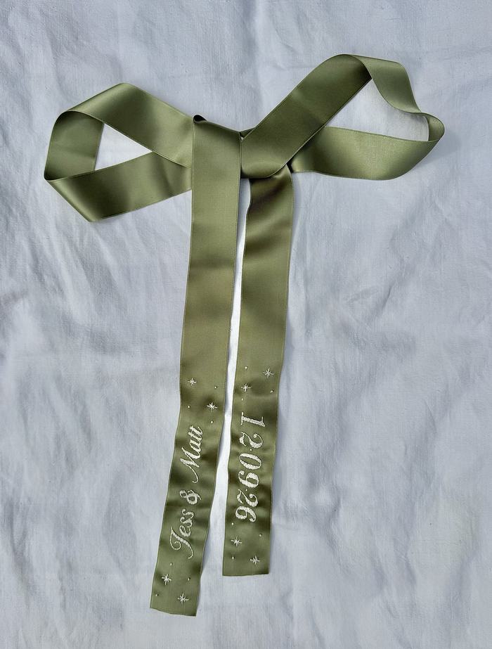 Sage green satin bow ribbon embroidered with names, a date, and star motifs